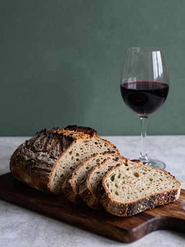 Artisanal sourdough bread sliced on a dark wood board, a glass of natural red wine beside it, moody side lighting, high contrast shadows.