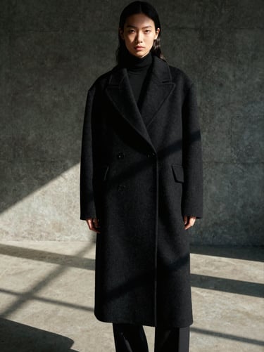 Editorial portrait of a high-fashion model wearing a structured black wool coat, standing in a minimalist concrete space, harsh side sunlight casting deep shadows, chiaroscuro, 35mm film grain.
