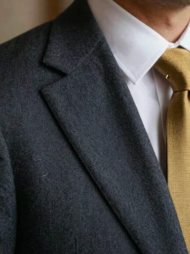 Close-up of a sharp charcoal grey suit shoulder, fine wool texture, crisp white collar, gold tie bar.