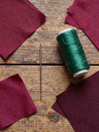 A flat lay photograph of sewing tools—a vintage needle, forest green thread, and dark magenta fabric scraps—arranged on a rustic wooden table in a whimsical workshop, Western / Global.