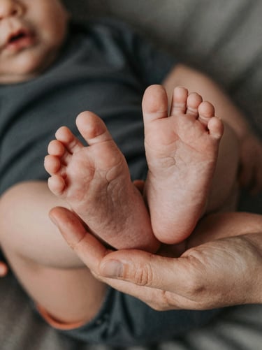 Details of a baby's tiny feet being held by a parent's hand. Anthracite and terracotta color accents in the clothing. Soft cinematic lighting, intimate and emotional atmosphere, European / French style.