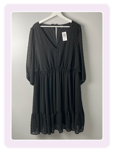 Black long-sleeve Swiss dot mini dress with a V-neckline hanging on a white hanger against a gray wall.