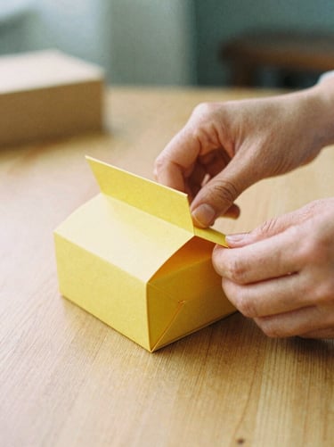 Close-up of artisan hands carefully folding a structured saffron-colored paper box on a clean wooden table, soft diffused morning light, 35mm film