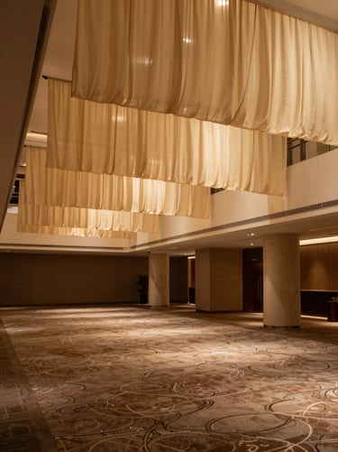 Finished hotel atrium interior photographed wide under warm tungsten light — fabric drapes, patterned carpet, and textured wall covering visible together, no human subjects, materials as the focal story