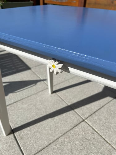 White daisy tablecloth weight clipped to a blue patio table on a sunny outdoor terrace.