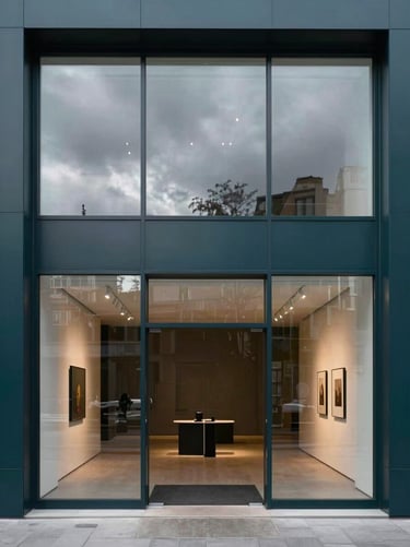 A portrait-oriented photography shot of a sleek, modern Northern European / Welsh / British gallery entrance with a large glass window reflecting a cloudy sky, showcasing a minimalist interior with deep charcoal teal accents.