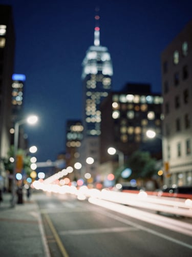 Artistic photography of motion-blurred lights in a North American / US cityscape at night. The dominant colors are deep navy blue and sharp off-white, creating a sense of energy and transformation. The composition is abstract, focusing on the atmosphere of a bustling metropolis.