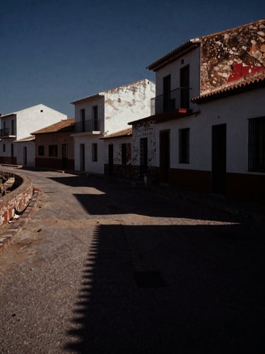 Dark cinematic film photography of a sun-bleached deserted Spanish coastal village, harsh midday sun casting long black shadows, eerie atmosphere, 35mm grain.