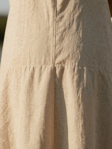 Extreme close-up of hand-finished seams on a beige linen dress, soft diffused side-lit daylight.