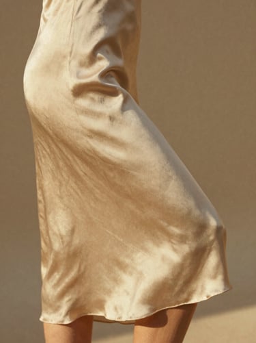 Detail shot of a soft gold silk dress skirt catching the light as it turns, warm low-contrast lighting.