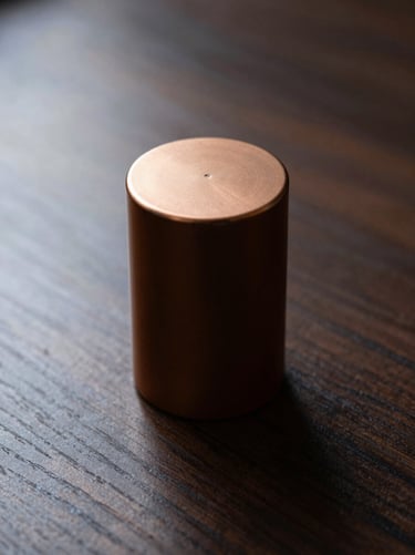 Close-up of a cylindrical perfume bottle with a polished bronze-toned cap resting on a dark wooden desk surface, raking light from upper left tracing the cap's edge, deep shadow behind bottle, sparse background