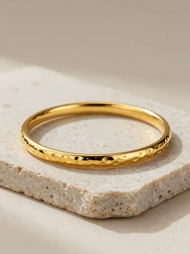 Three-quarter overhead product shot of a heavy 22-karat gold bangle pair on a smooth pale stone slab, soft warm studio fill light from the left, the bangles' hammered surface texture clearly visible, no hands or model, neutral ivory background