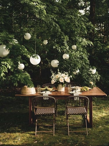 Shaded garden seating spot with wooden dining table, soft hanging lights and surrounding greenery fo