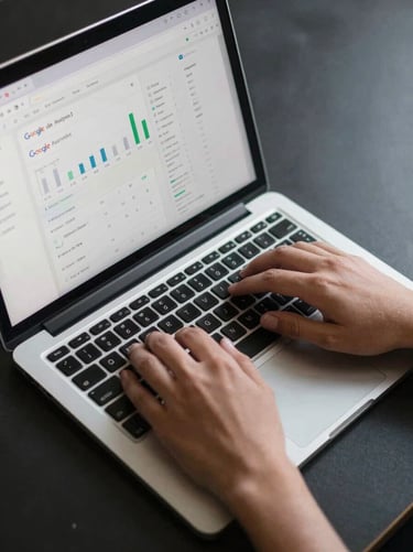 Overhead close-up of hands typing on a laptop keyboard, a browser tab showing a Google Analytics dashboard visible on screen, cool even studio lighting, minimal dark desk surface