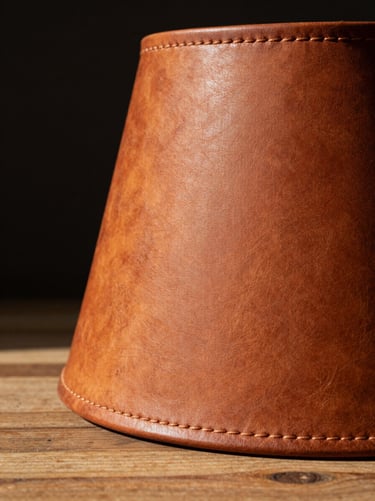 Extreme close-up, straight-on product study: a hand-stitched leather lamp shade resting on a raw wood surface, north-facing studio daylight raking across the surface to reveal every seam and hand-burnished ridge. The leather is warm amber-brown, the stitching thread visible and slightly irregular — proof of individual hand-work. Deep shadow fills the left third of the frame; the lamp's silhouette is crisp against a dark void background. No lifestyle props, no office setting.