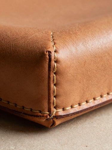 Macro detail shot of a leather lamp's hand-stitched seam, photographed straight-on under soft north-facing daylight. The thread is waxed and slightly uneven — each stitch interval slightly different from the last. The leather surface shows hand-burnishing marks along the edge, the patina deepening where fingers pressed. No background clutter; a plain warm linen surface beneath. No lifestyle props, no person visible beyond the finished seam.