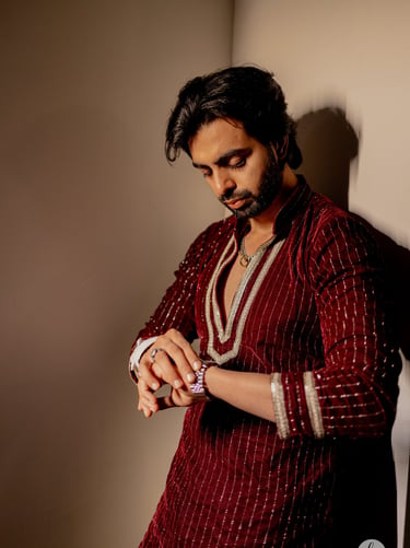A man in a maroon embroidered Indian kurta checking his wristwatch while leaning against a wall.