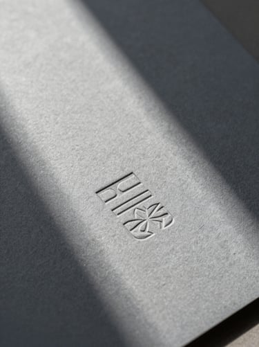 Extreme close-up of embossed stationery on heavy cool ash gray paper stock, hard directional natural light casting a sharp diagonal shadow across the texture.