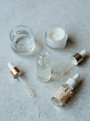 Overhead composition of natural skincare ingredients and a clean glass dropper on a warm light gray stone surface, soft diffused light.