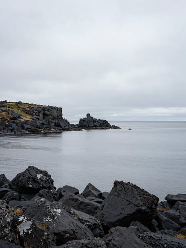 A wide-angle landscape photograph of a rugged Northern European coast, featuring dark charcoal volcanic rocks meeting a calm, light gray sea under a misty sky. The composition is minimalist and serene, emphasizing vast, quiet space.