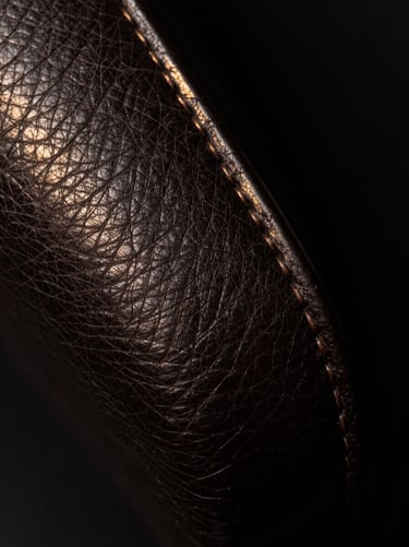 Extreme close-up of hand-stitched dark leather grain, catching a faint golden light against absolute black shadows