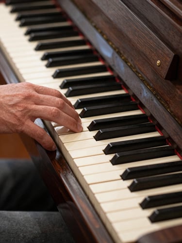 A detailed close-up of a hand resting on the keys of an antique upright piano in a North American / US music hall. The image has a timeless, elegant feel. The keys are a soft pale cream and the wood is a rich dark charcoal brown.