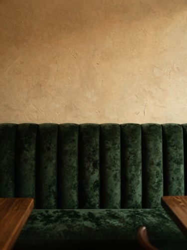 A quiet corner of the restaurant, dark green velvet banquette seating against a textured warm ivory wall, soft shadows