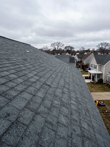Wide environmental shot of a completed residential roof replacement in a DC-area neighborhood, overcast daylight, full roofline visible from a slight elevation angle showing clean architectural shingle lines and ridge cap, no people