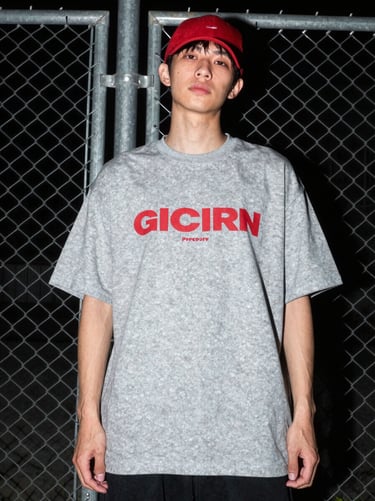 Low-angle gritty streetwear photo of a model wearing an oversized grey graphic tee, direct flash, posing against a metal chain-link fence at night