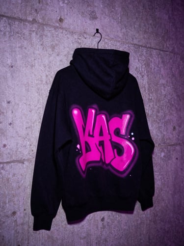 Close-up of a premium heavy cotton black streetwear hoodie with a vibrant magenta graffiti print on the back, hanging on a raw concrete wall.