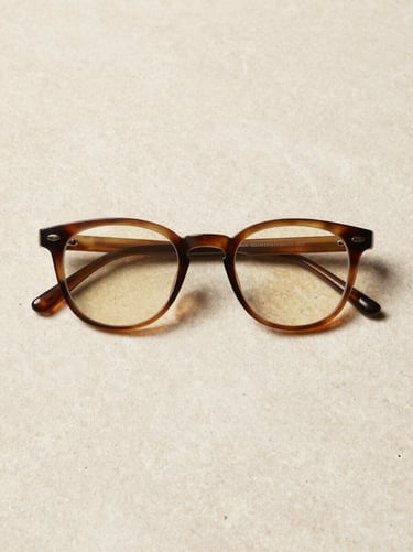 High-premium designer eyewear frame resting flat on a honed limestone surface, overhead flat-lay composition, controlled north-facing studio light, subtle umber shadow at frame edges, warm cream stone texture, no lifestyle context, clean product isolation