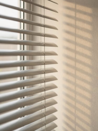 Angled close-up of aluminium venetian blind slats, fine horizontal lines casting parallel shadow bands on a warm cream wall, soft diffused daylight, no people