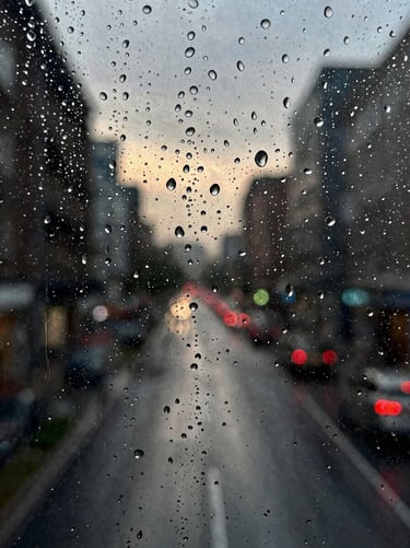 A close-up of rain droplets on a textured glass pane, with the soft-focus silhouette of a city street in the background at dusk.