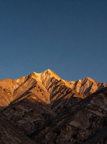 A breathtaking cinematic shot of the Atlas Mountains at dusk, with the peaks glowing in soft gold light under a deep indigo North African / Moroccan sky.