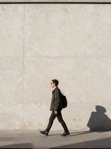 A minimalist street photography shot in New York City. A lone figure in contemporary attire walks past a clean, light-colored concrete wall with sharp shadows. Bright, direct North American afternoon sunlight. Quiet, artistic composition.