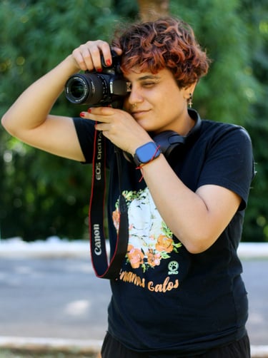 A young, short-haired person with curly brown hair uses a Canon DSLR camera with a black lens to take a photo outdoors