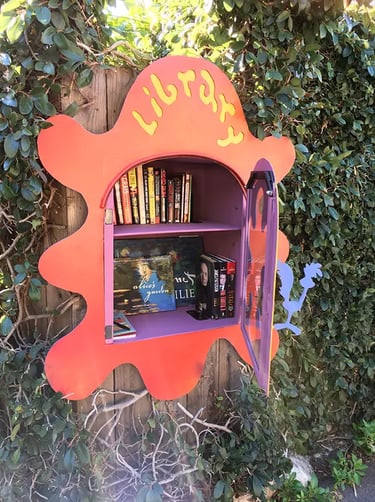 An red, splat-shaped Little Free Library box mounted on a wooden fence, filled with books and a purple open door.