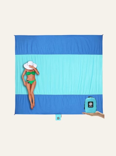 Woman in a green bikini lying on a large sand-free beach blanket with its compact carrying pouch.