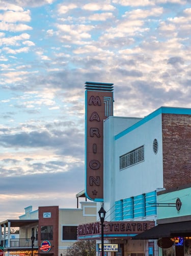 Exterior of the Marion Theatre building under a partly cloudy sky, featuring its tall vertical neon 