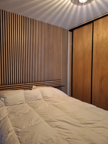 Lit blanc avec panneau en latte de bois clair servant de tête de lit luminaire noir et mobilier bois