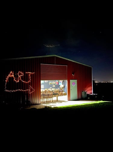 Red barn art studio at night with fairy lights and an open door showing interior paintings.