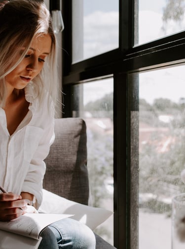 Woman in a white linen shirt writing in a notebook by a bright window.