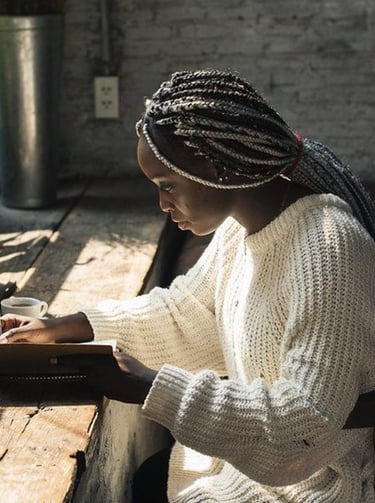 Young woman writing in a journal at a rustic wooden desk by a sunny window with books.