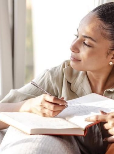 A young woman writing in her journal while looking out a window thoughtfully.
