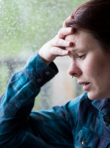 A stressed woman rests her head on her hand near a rainy window, depicting anxiety and mental health struggles.
