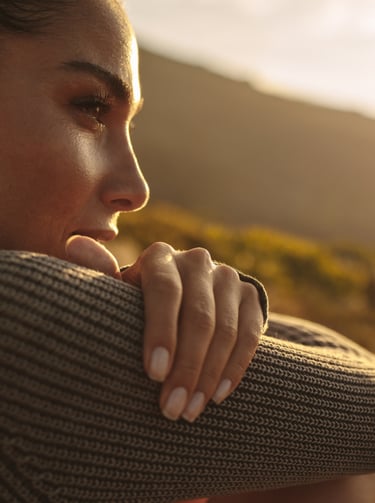 A young female hiker in a grey sweater looks at the golden sunset over mountain hills.