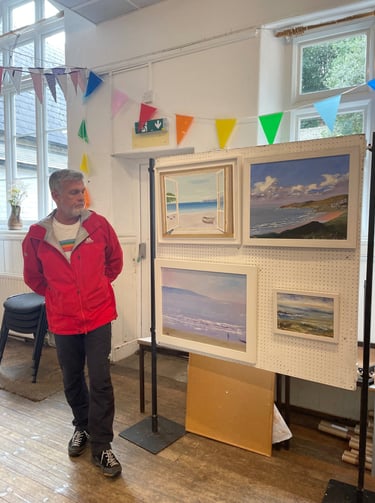 Artist Chris Gill standing beside framed coastal oil paintings at New Atlantic Wave Art Exhibition, 