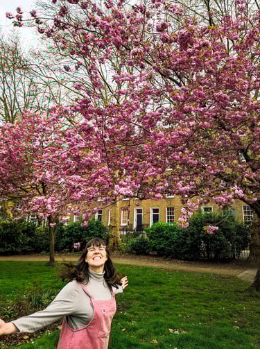 Laura dances in front of a pink blossom tree in London. she is wearing pink overalls and smiling.