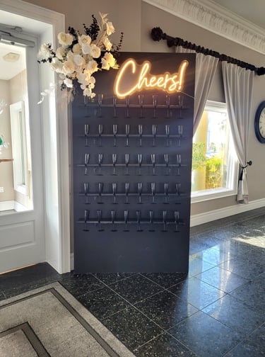Matte black champagne wall with black champagne glasses, white floral arrangement and a cheers neon