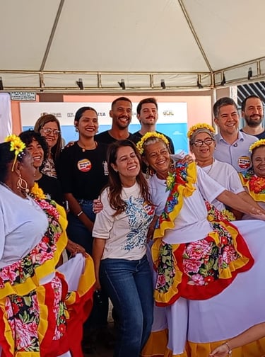 A group celebrates at a cultural event with women in traditional floral Baiana dresses and local community leaders.
